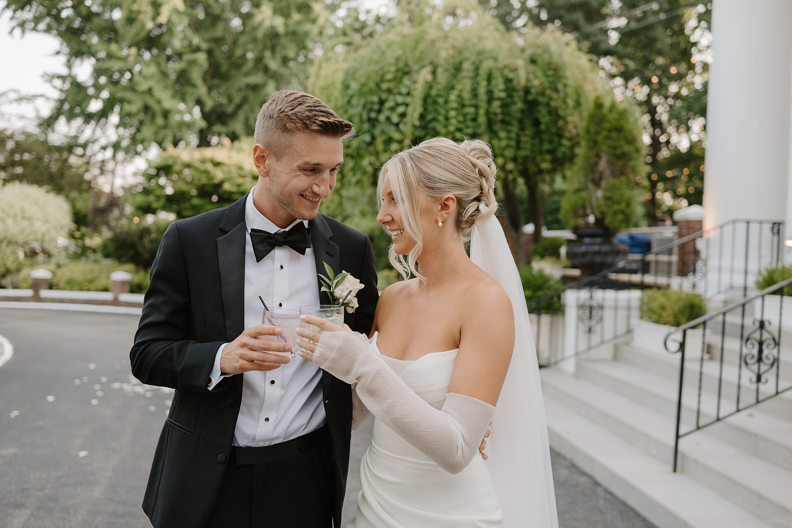 Bride and groom smile at each other as they clink their signature cocktail glasses outdoors.